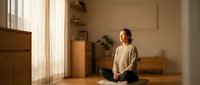Young Asian woman practicing mindfulness meditation in minimalist home interior with natural sunlight streaming through window creating peaceful atmosphere.