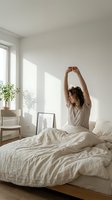 Woman with dark hair and medium skin tone stretching in bed after waking, wearing casual beige shirt in bright, minimalist bedroom with natural sunlight for wellness and lifestyle content.