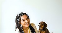 Young woman with braided hairstyle and beads posing with small brown dachshund dog against white background for pet care and lifestyle content.