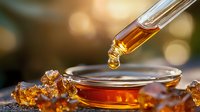 Amber liquid drop falling from dropper into glass dish with golden bokeh background, macro photography capturing honey-like substance in motion.