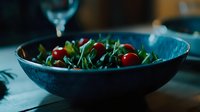 Fresh arugula salad with cherry tomatoes in dark ceramic bowl against moody teal background, dramatic food photography with natural lighting.