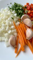 Fresh chopped vegetables including diced onions, carrots, red bell peppers, garlic cloves and herbs prepared for cooking on white cutting board.