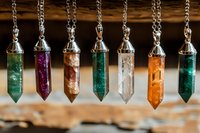 Crystal pendants in various colors hanging on silver chains against wooden background, featuring emerald, amethyst, quartz and amber tones for spiritual jewelry.