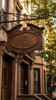 Vintage wooden hanging sign with ornate carved frame mounted on historic brownstone building facade with decorative ironwork and lush green foliage.