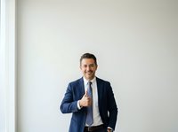 Confident male professional in navy blazer and light shirt giving thumbs up gesture against white wall background, studio portrait for corporate and business communications.