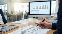 Business professionals analyzing financial data and charts in modern office meeting room with computer monitor displaying growth statistics.