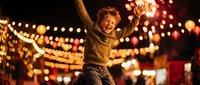 Young blonde woman celebrating with arms raised at night festival with warm bokeh lights and string illumination creating joyful atmosphere.