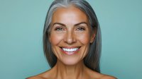 Confident mature woman with long gray hair and natural makeup smiling warmly at camera against turquoise background, representing aging gracefully and self-acceptance.