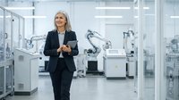 Senior woman in dark business suit walking through modern industrial manufacturing facility with robotic automation equipment and machinery in background.