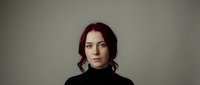 Professional portrait of confident young Caucasian woman with burgundy hair wearing black turtleneck against neutral background for business branding.