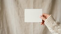 Person with medium skin tone wearing a cream knit sweater holds a blank white card against a neutral beige fabric background, suitable for mockups and promotional designs.