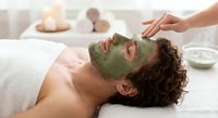 Young Caucasian man receiving green clay facial mask treatment at luxury spa with therapist hands applying skincare for relaxation and wellness.