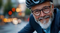 Mature bearded man in cycling gear and glasses smiling at camera with blurred city lights in background, expressing active lifestyle and vitality in senior years.