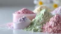 Colorful pastel powders in pink, mint green, and purple shades arranged in piles and measuring cups on white surface, soft focus macro photography with daisies in background.
