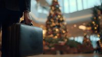 Business traveler with suitcase at airport terminal during Christmas holiday season with decorated tree and warm bokeh lights in background.