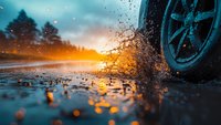 Car wheel splashing through puddle on wet road during dramatic sunset, creating dynamic water spray with golden light bokeh reflections and silhouetted forest background.