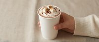 Hand holding a white paper cup of hot chocolate topped with whipped cream and marshmallows on a beige linen background.