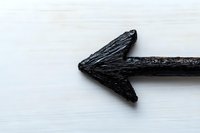Black charcoal arrow pointing left on white wooden background, minimalist composition showing direction and guidance for business concepts and design.