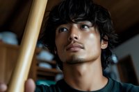 Young Asian male with wavy black hair and thoughtful expression looking up, wearing dark t-shirt against wooden interior background in natural lighting.