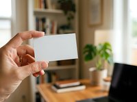 Hand holding blank white business card in modern home office with bookshelf, laptop, and plants, suitable for branding and corporate identity mockups.