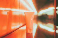 Coral-colored cosmetic tube on reflective surface with blurred neon lights creating dynamic background atmosphere, suitable for beauty product advertising.