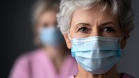 Senior Caucasian woman wearing protective surgical face mask during virus pandemic, with healthcare worker in background, dark moody lighting.