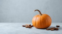 Orange pumpkin with cinnamon sticks and star anise spices on white table against gray background for autumn seasonal cooking and decoration.