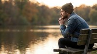 Young man in denim jacket and beanie sitting on wooden dock by lake during golden hour sunset, contemplating peacefully in autumn nature setting.