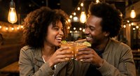 Happy African American couple toasting with beer glasses in cozy restaurant with warm string lights creating romantic dining atmosphere.