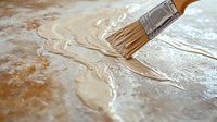 Paint brush applying smooth beige acrylic paint strokes on textured canvas surface, creating flowing abstract artistic pattern with metallic sheen effect.