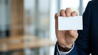 Mature male professional in dark business suit holding blank white business card in office environment with blurred background for corporate branding.