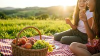 Two young women enjoying a sunny outdoor picnic with a basket of fresh fruits on a red checkered blanket in a green meadow.