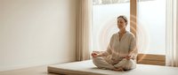 Young Asian woman in beige loungewear practicing meditation in lotus position on yoga mat by bright window in minimalist home interior for wellness.