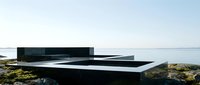 Minimalist black architectural structure on rocky coastal landscape with calm water and clear sky in background.