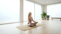 Young blonde woman practicing meditation in lotus pose on yoga mat in bright modern room with large windows and plants for wellness lifestyle.