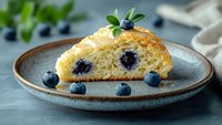 Fresh baked blueberry scone with golden crust served on ceramic plate, garnished with fresh berries and mint leaves, close up view on blue background.