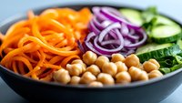 Fresh buddha bowl with spiralized carrots, chickpeas, red onion rings and sliced cucumber in dark ceramic bowl, healthy vegetarian meal prep.