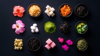 Colorful Asian tea ceremony ingredients and snacks arranged in neat rows on dark background, including flowers, sugar cubes, matcha powder and dried tea leaves.