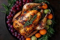 Roasted whole chicken with crispy golden skin served on dark plate with fresh grapes, oranges, brussels sprouts and aromatic herbs for festive dinner.