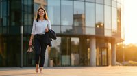 Confident young Caucasian businesswoman walking barefoot outdoors near modern glass office building at sunset, holding jacket and heels.