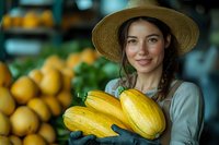 Young Caucasian woman in straw hat holding fresh yellow squash at farmers market, genuine smile, wearing casual attire against blurred produce background.