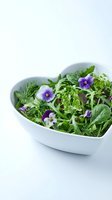 Fresh mixed greens and edible flowers in heart-shaped white bowl on white background. Healthy salad with purple pansies, microgreens, and herbs for food styling and nutrition content.