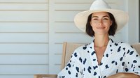 Young Caucasian woman in white sun hat and polka dot dress smiling while relaxing on porch chair, expressing joy and contentment during summer leisure time.