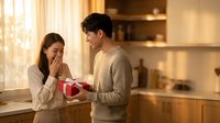 Young Asian couple sharing romantic moment in modern kitchen as man presents gift box to surprised woman during golden hour lighting for Valentine's Day celebration.