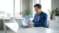 Young Caucasian businessman in blue shirt working on laptop at modern office desk with plants, analyzing financial data charts for business growth strategy.