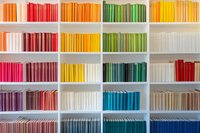 Colorful books arranged by spectrum on white shelves create rainbow pattern. Organized library collection shows spines in gradient from warm to cool tones. Modern minimalist bookshelf display.