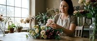 Young Asian woman florist arranging fresh flower bouquet at wooden table in bright workshop studio with natural light from large windows.