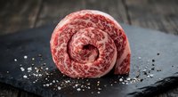 Raw marbled beef roll tied with string on dark slate surface with coarse salt and peppercorns for gourmet cooking and culinary preparation.