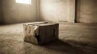 Old damaged cardboard box sitting alone on concrete floor in empty warehouse room with natural window light creating atmospheric shadows.