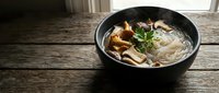 Asian noodle soup with mushrooms and herbs in black bowl on rustic wooden table for restaurant menu design and culinary content.